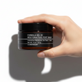 A hand holding a jar of Formula No. 32 Restorative Foot Balm against a white background with cinematic shadows.